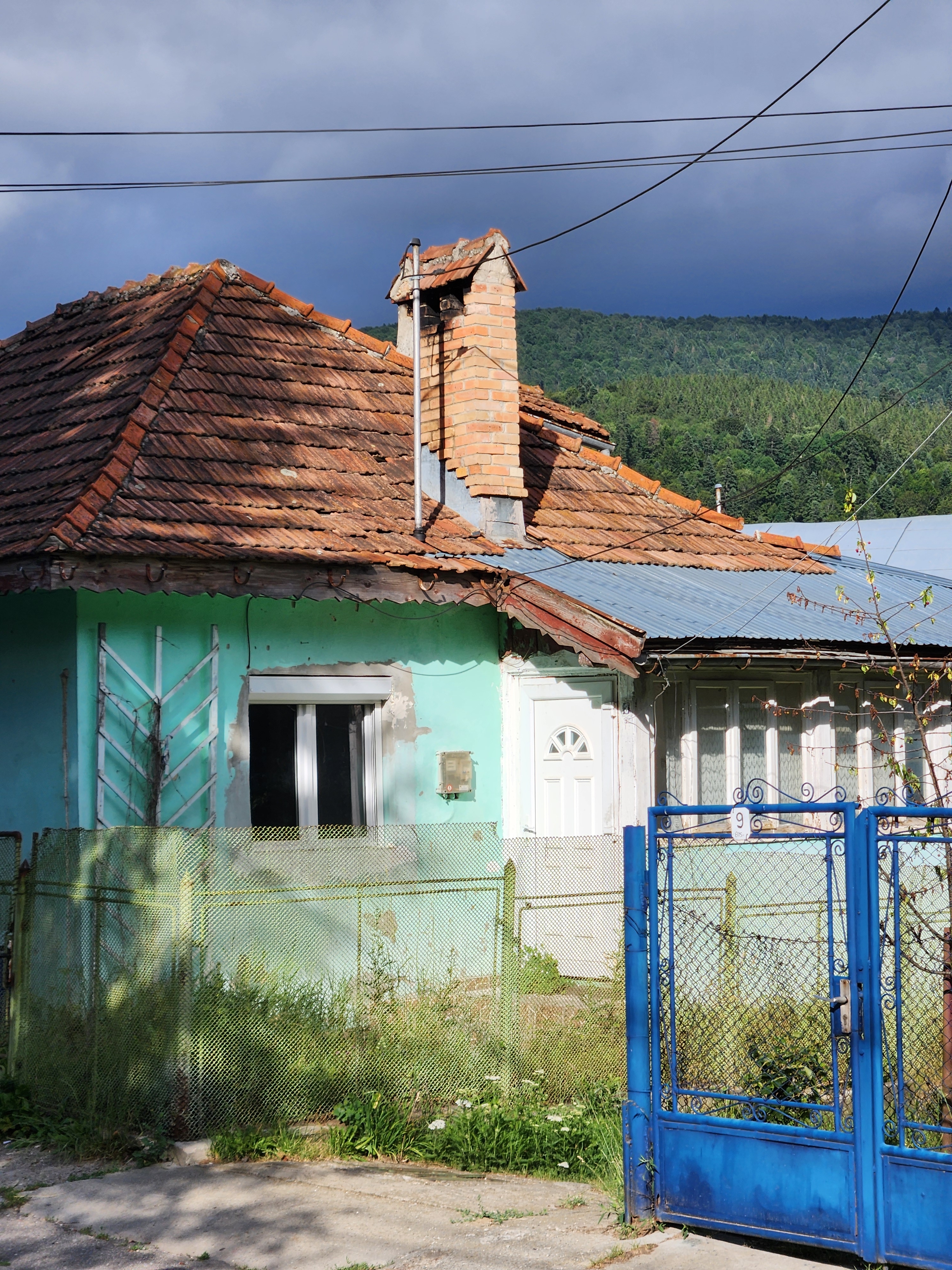 A house painted turquoise, in bright sunlight half blocked by the dark shadow of a tree, with a backdrop of a heavy grey sky and forested mountains