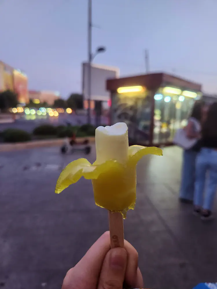 A hand holding a banana shaped ice cream up against a city street at dusk