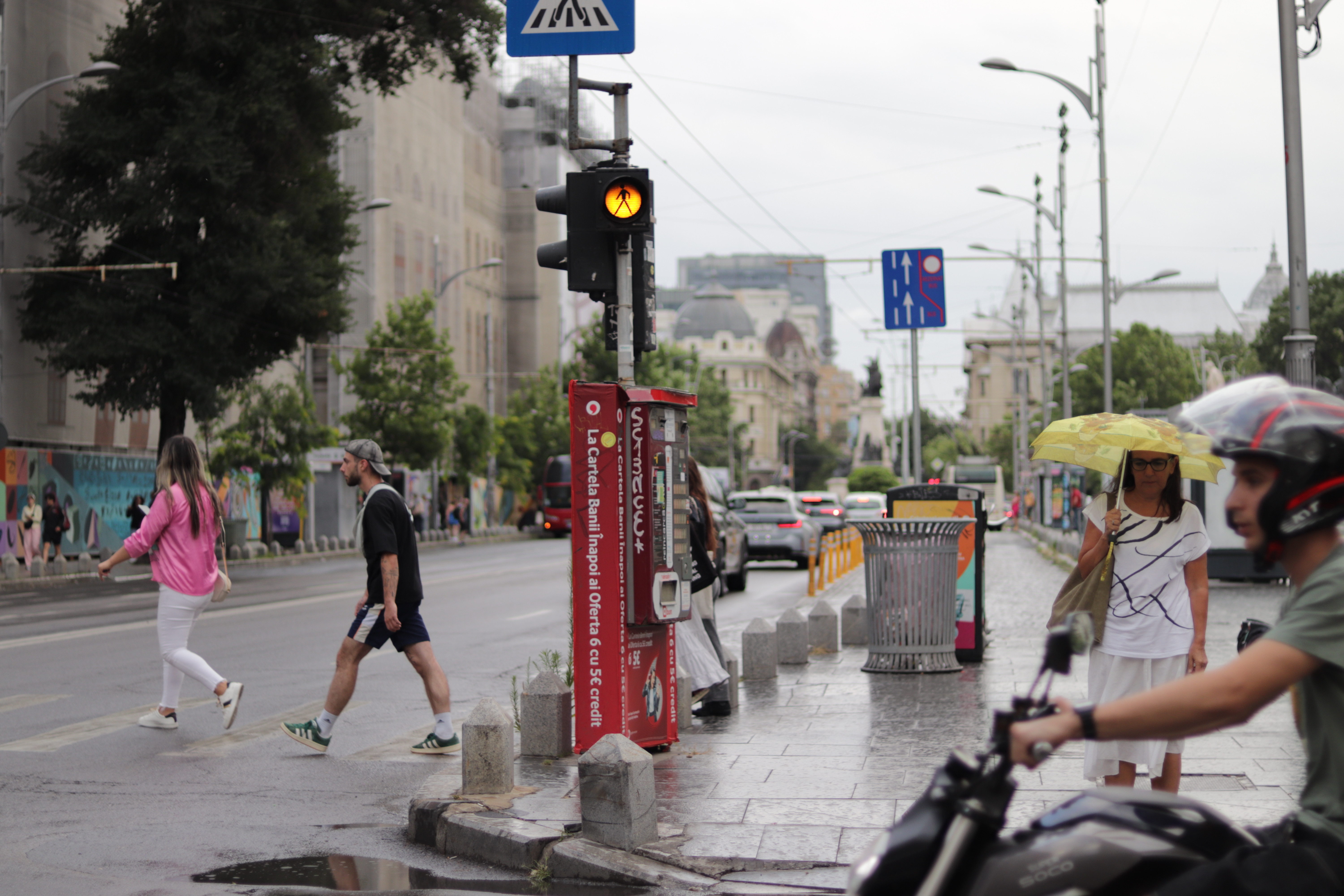 a red kiosk by a street crossing with people walking past in two directions, and a motorcyclist waiting for a traffic light in the foreground