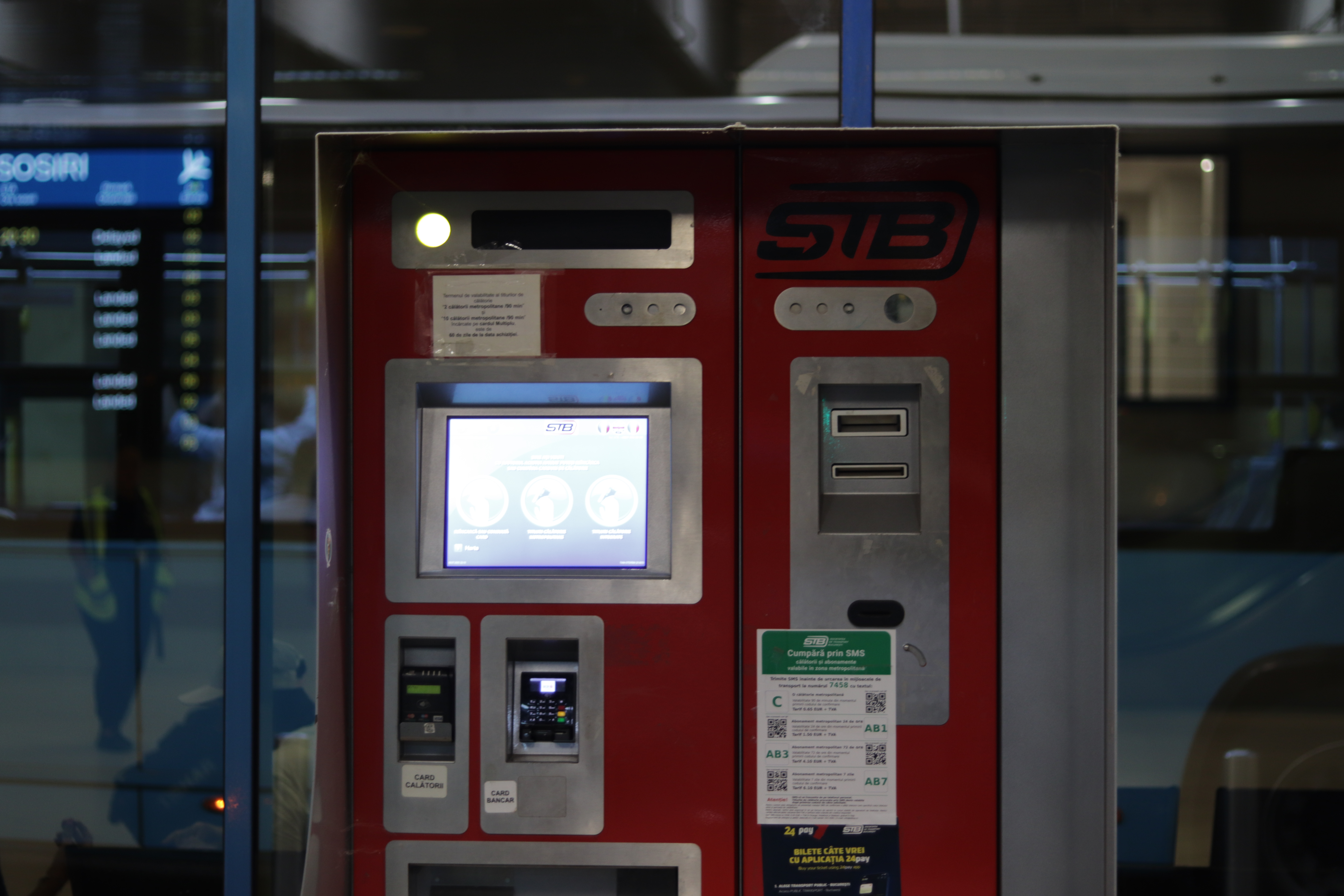 a red metal kiosk in front of a glass wall through which an airport departures board can be seen