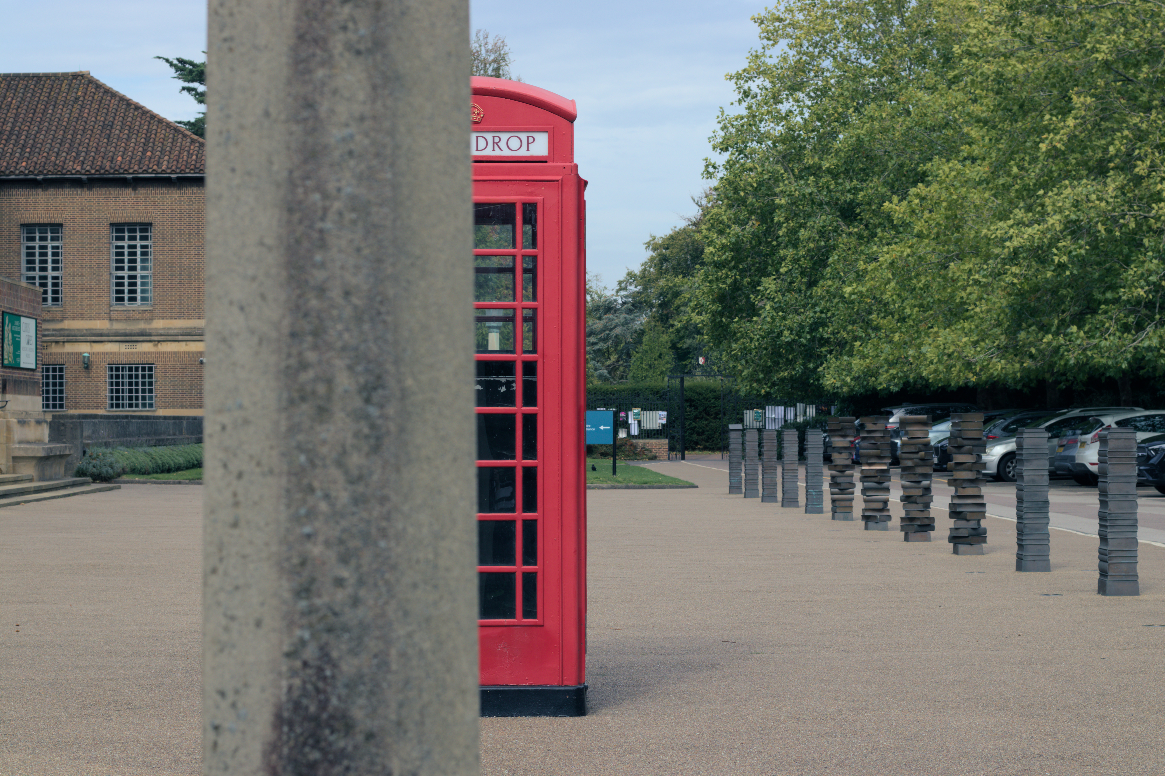 A red phone box half obscured by an out of focus concrete pillar, in the background are a library and bollards made to look like stacks of books