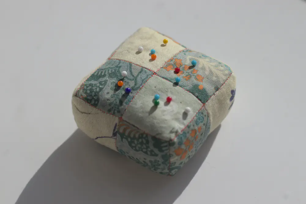 a green cuboid pincushion with multicoloured pins stuck into it randomly