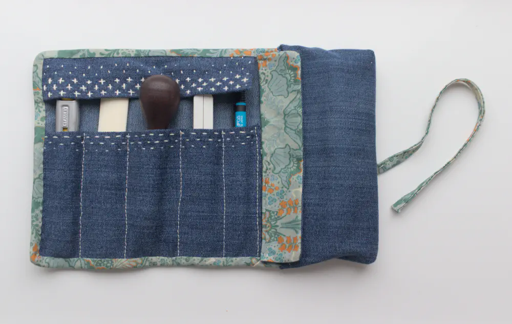 a denim tool roll with green patterned edging and white sashiko decoration, holding tools such as a craft knife, bone folder, awl, scissors and mechanical pencil