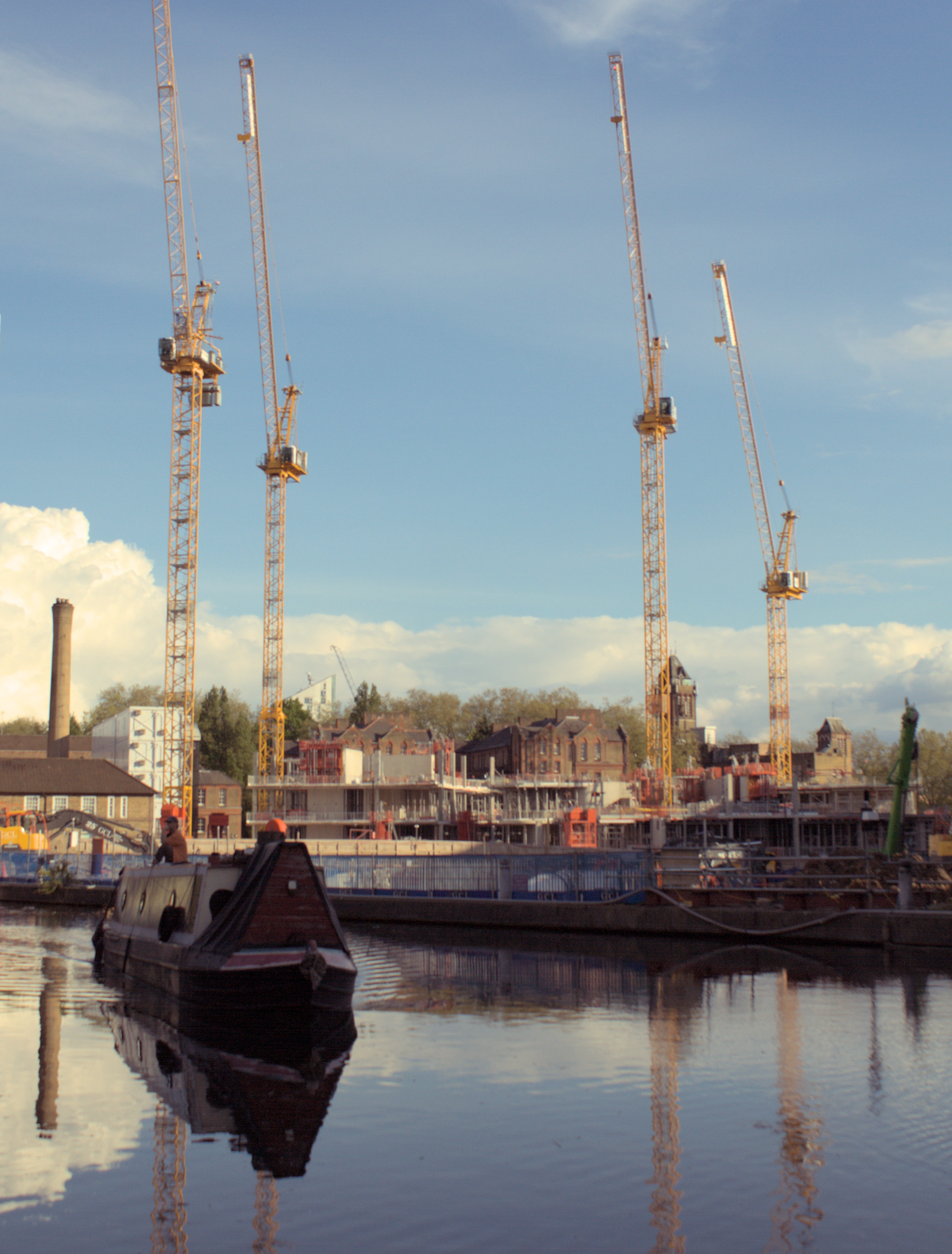 A boat on a canal with 4 warm yellow cranes rising from a construction site behind it