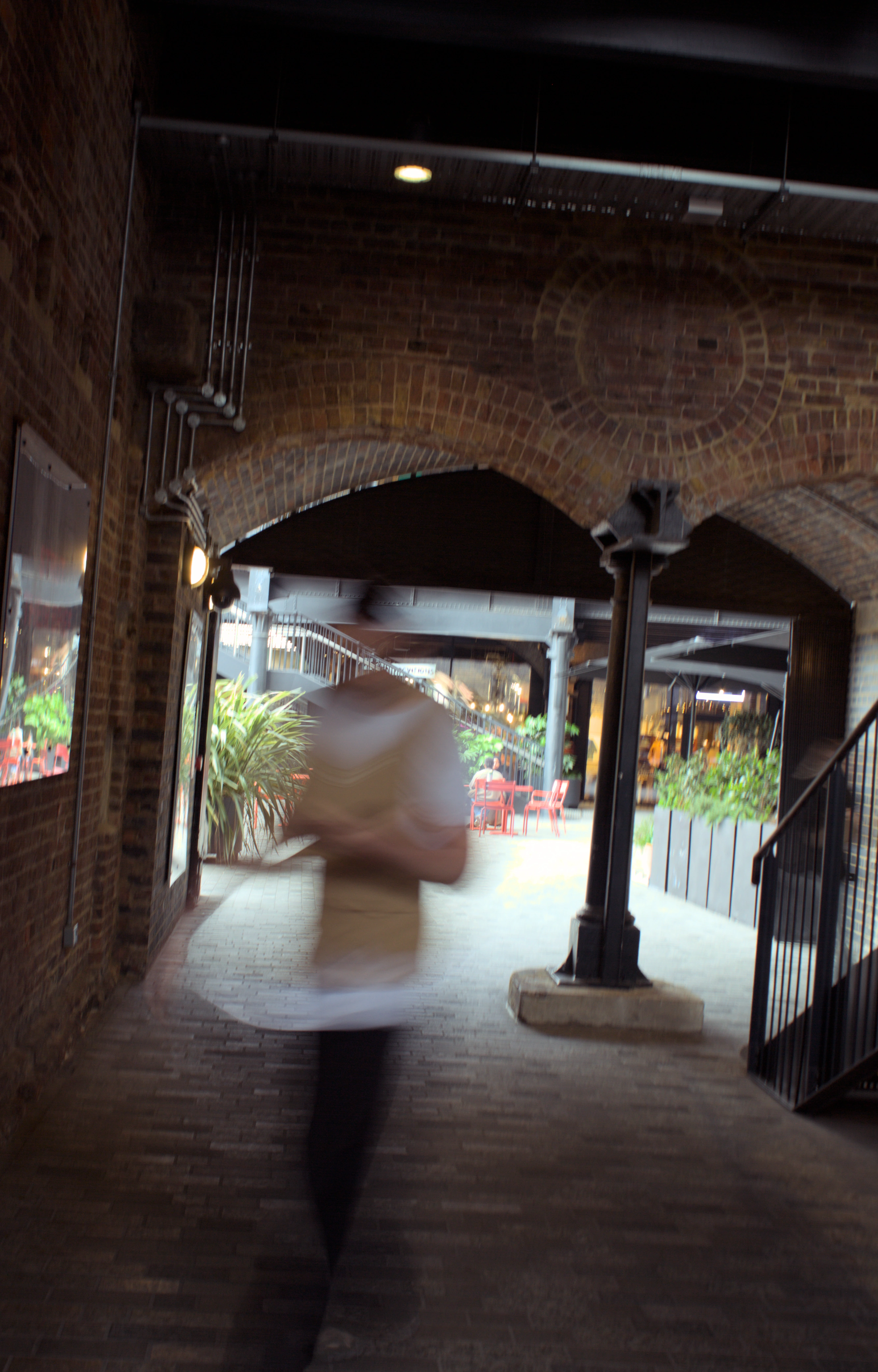 A blurry figure in front of an archway with green plants and red chairs beyond