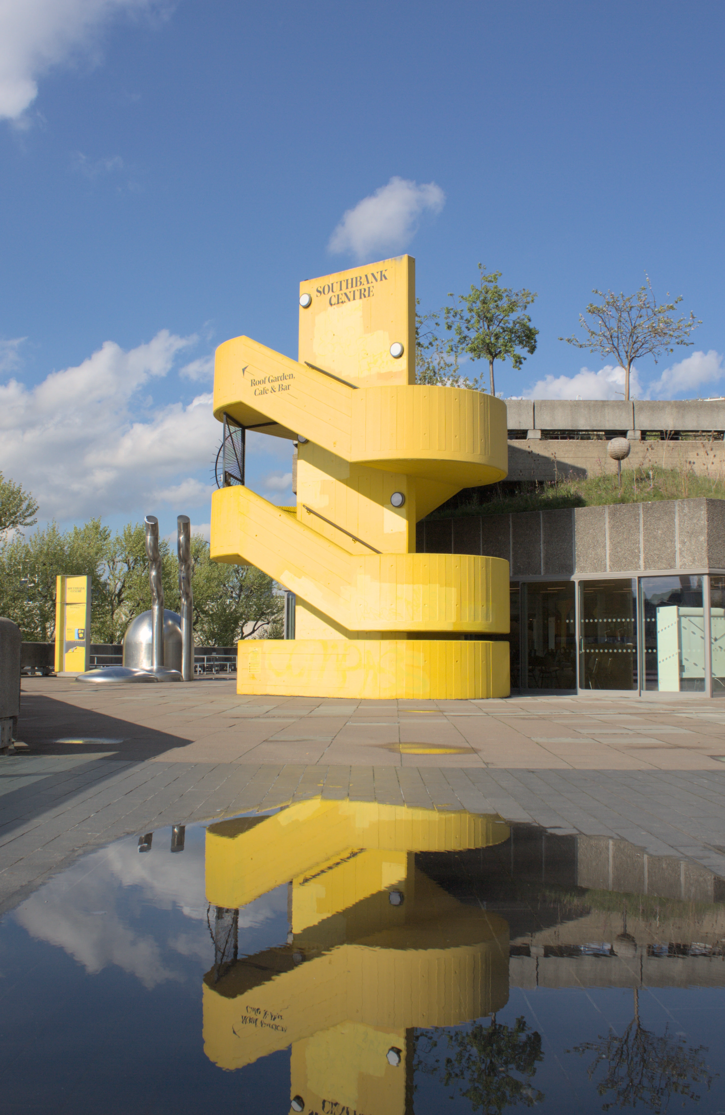 A bright yellow brutalist staircase against a blue sky, reflected in a puddle below